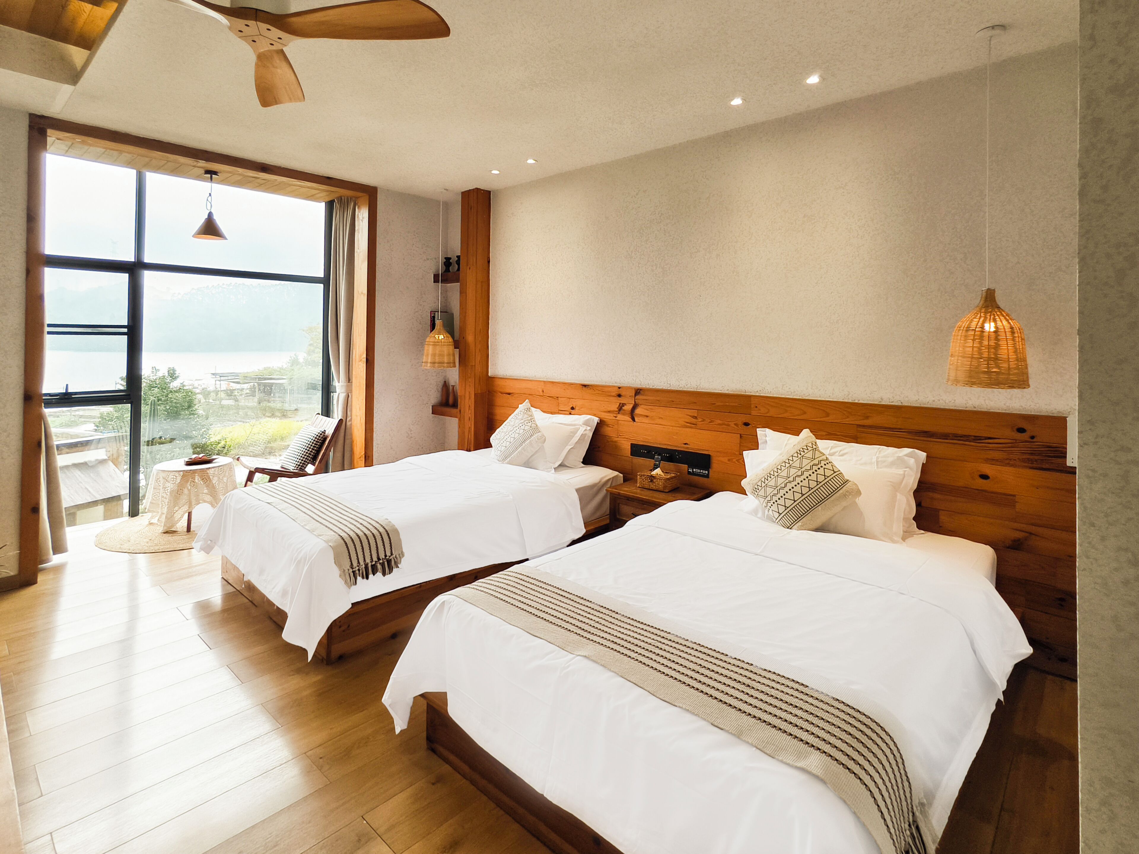 Une chambre d’hôtel avec deux lits, une grande fenêtre donnant sur un beau paysage, un plancher en bois et un ventilateur de plafond.
