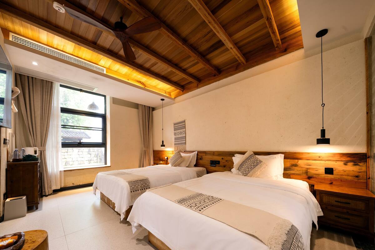 Ein Hotelzimmer mit zwei Betten, Holz-Kopfteilen, einem großen Fenster mit Vorhängen, einer Holzbalkendecke und einer Hängeleuchte.