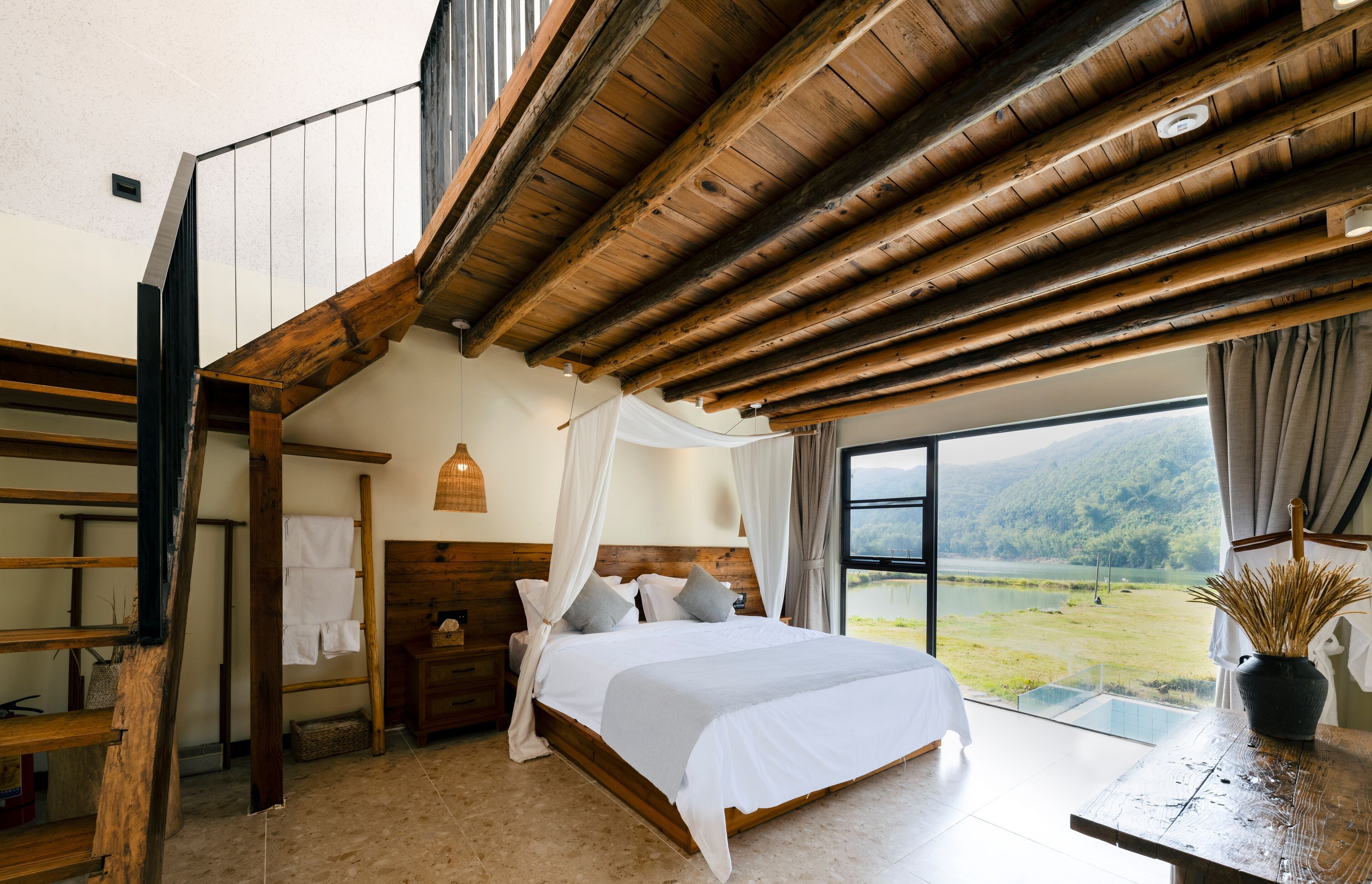 Un dormitorio con techo de madera, una cama grande con ropa blanca, una mesita de noche y vistas a un lago y montañas.