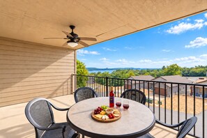 Une terrasse extérieure couverte, avec une table ronde, des chaises, une bouteille de vin et une vue sur un lac et des arbres.