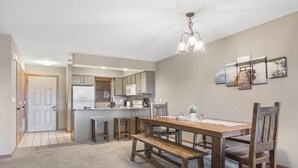 Un coin repas avec une table et des bancs en bois, une cuisine à l’arrière-plan et une porte donnant sur une autre pièce.