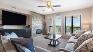 A living room with a flat-screen TV, dark wooden cabinets, a stone accent wall, a ceiling fan, and a view through the glass doors. - 2 BD Lake View Condo, with Jacuzzi Tub, Covered Deck, Resort Amenities and Discount Tickets (Branson)