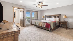 A bedroom with a bed, bedside lamps, a ceiling fan, a wooden desk, and a door leading to a balcony.