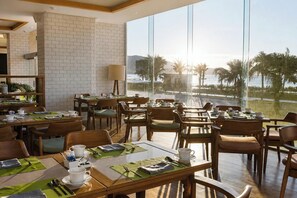 A dining area with multiple tables and chairs, a brick wall, and a large window with a view of palm trees and a body of water.