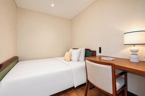 A hotel room with a single bed, a bedside table, a chair, and a desk with a lamp.
