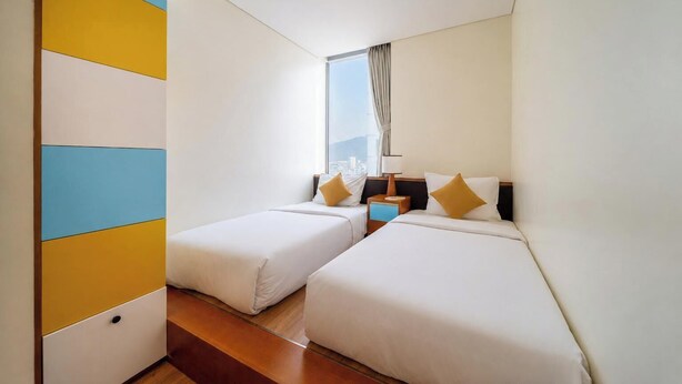 A hotel room with two beds, a window with curtains, a nightstand, and a view of the city. - Sea and Sun Apartments (Da Nang)