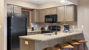 Une cuisine dotée de plans de travail en granit, d’un réfrigérateur noir, de meubles en bois, d’un four à micro-ondes, d’une cuisinière et d’un évier. On trouve trois tabourets en bois près de l’îlot central.