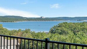A coastal view with a large body of water, a distant building on a hill, and dense green foliage in the foreground. - Sweet 3BR + Loft With Spectacular View of Lake (Branson)