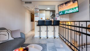Un salon moderne avec un canapé, une table basse sur laquelle sont disposés des fruits, et un coin cuisine comprenant un bar et des tabourets.