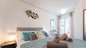 Un lit bien fait, avec une couvre-lit à motifs, des oreillers et des serviettes enroulées. On peut voir une table de chevet avec une lampe et un objet décoratif fixé au mur.