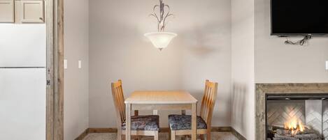 A dining area with a table and chairs, a hanging light fixture, a fireplace, and a television mounted on the wall.