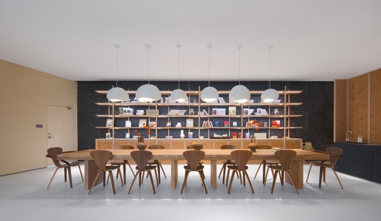Un comedor moderno con una mesa de madera larga y sillas, una estantería central e iluminación colgante.
