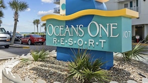A sign for Oceans One Resort with a modern design, featuring a wave motif and the resort's name. - Wake up to sweeping ocean views from the 16th floor at Oceans One (Myrtle Beach)