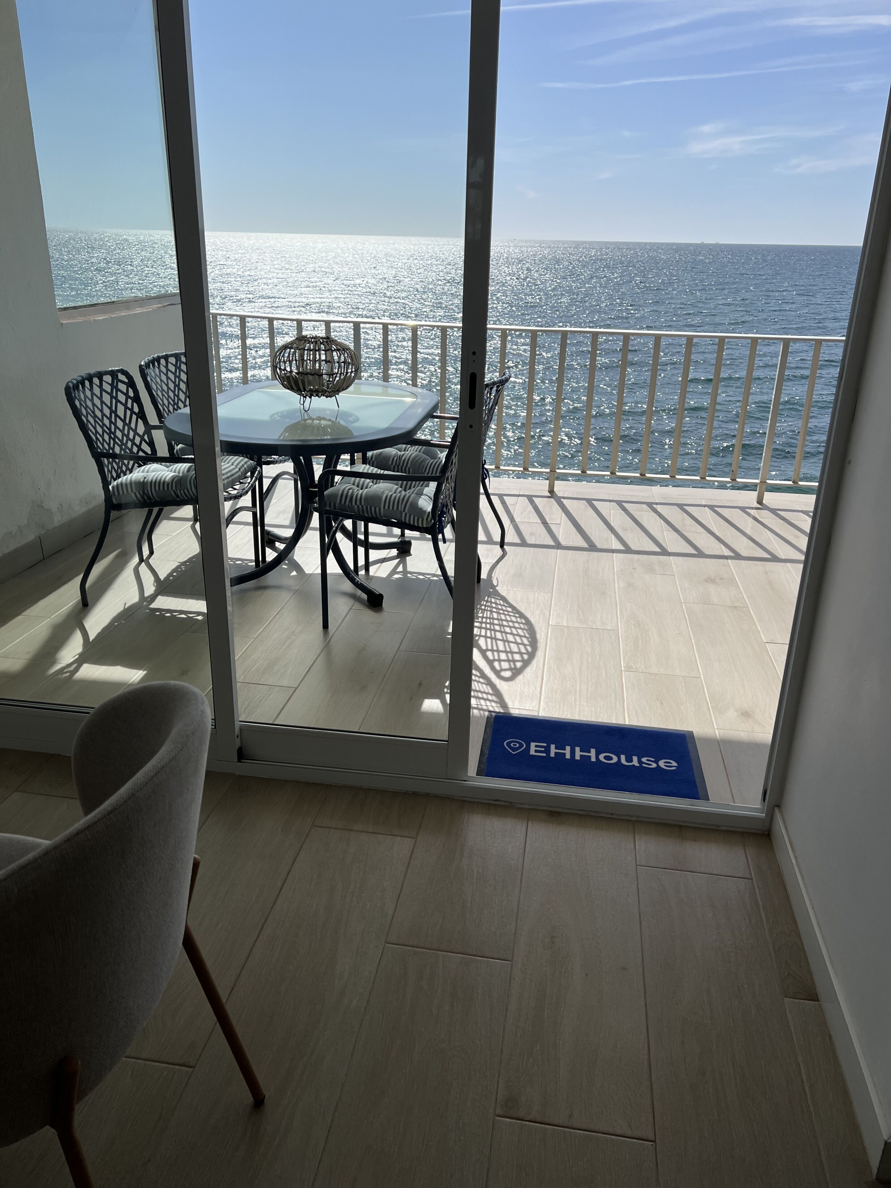 A balcony with a glass door leading to a terrace with a table and chairs, overlooking the sea.