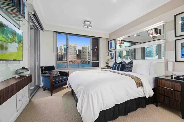 A modern hotel room with a large bed, a seating area with a blue sofa, a side table, and a view of the cityscape through large windows.