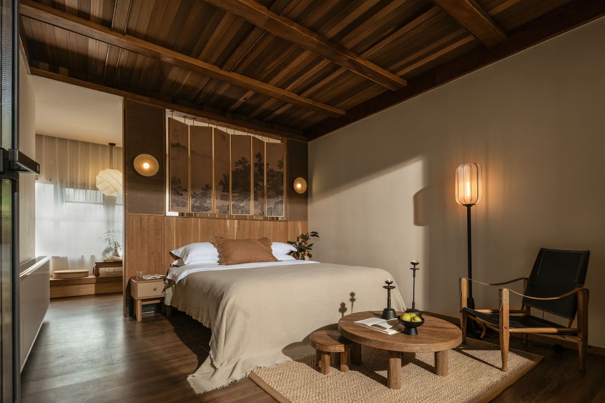 A bedroom with a wooden ceiling, a bed with a beige bedspread, a bedside table with a lamp, a chair, and a small round table with a book and a bowl of fruit.