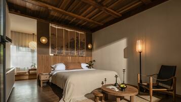 A bedroom with a wooden ceiling, a bed with a beige bedspread, a bedside table with a lamp, a chair, and a small round table with a book and a bowl of fruit.