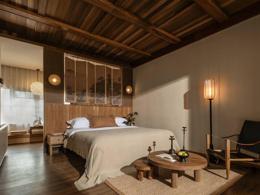 A bedroom with a wooden ceiling, a bed with a beige bedspread, a bedside table with a lamp, a chair, and a small round table with a book and a bowl of fruit.