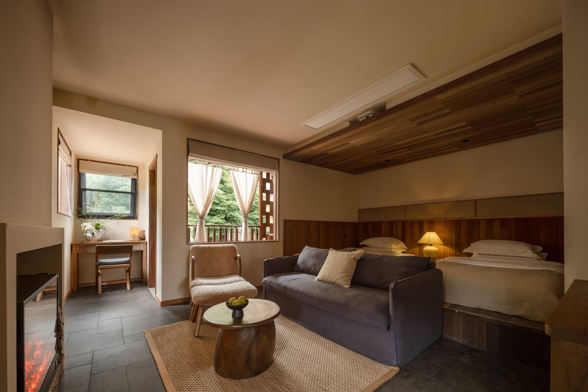 A cozy room with a sofa, a small table with a bowl of fruit, a chair, a desk, and a bed with a headboard.