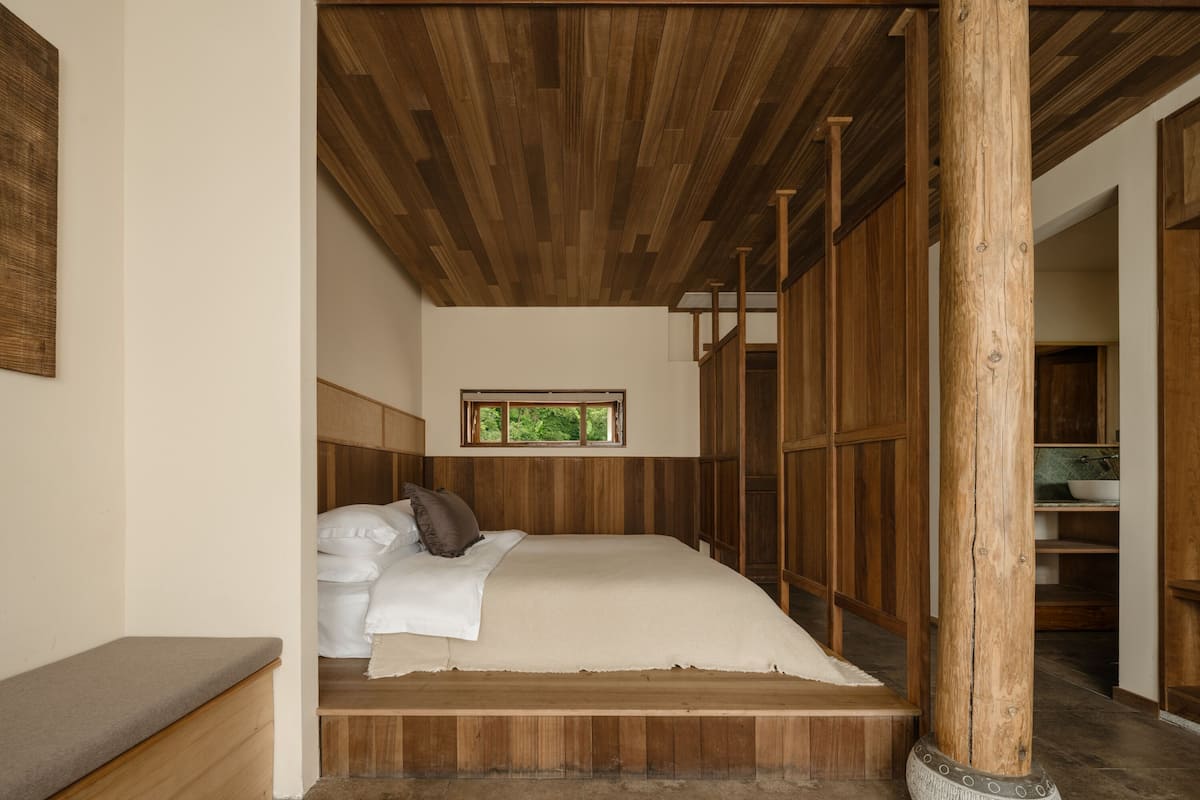 A wooden-framed bedroom with a bed, a bench, and a view of a kitchen area.