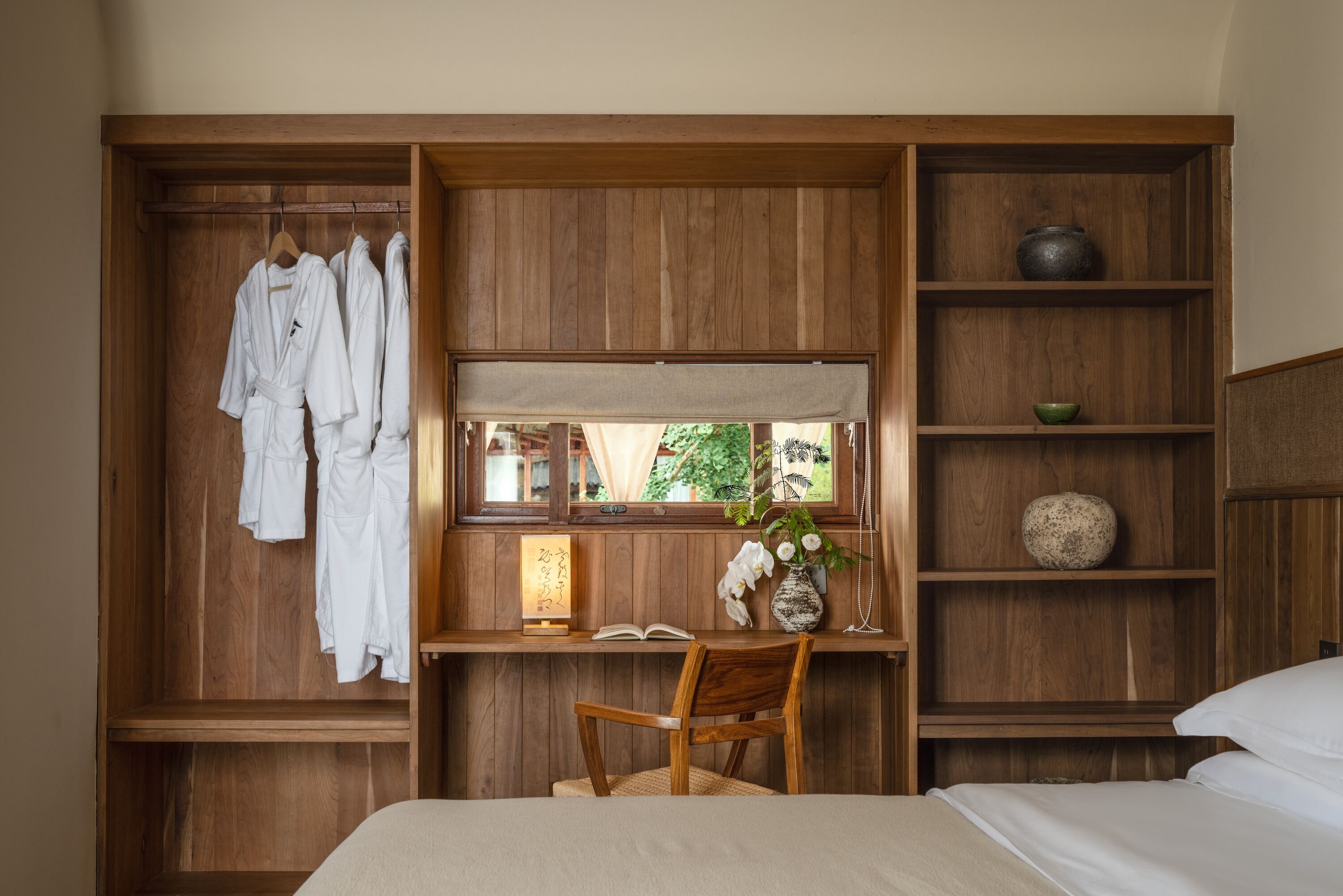 A room with a wooden wardrobe, a desk with a chair, a bed, and a window with a view of greenery.