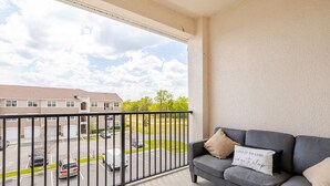 Un balcon avec un canapé gris, un coussin orné d’une citation inspirante, et une vue sur un parking et des immeubles.