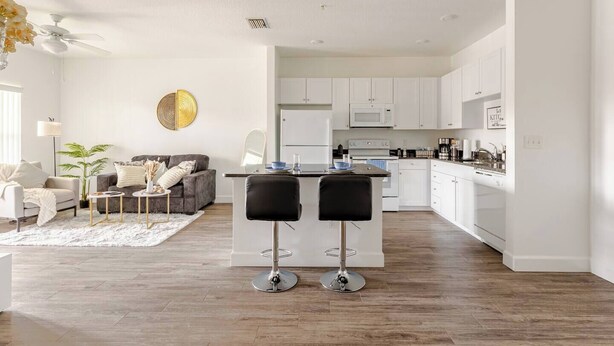 Une cuisine moderne avec des meubles blancs, un îlot central doté de deux tabourets et un coin repas comprenant un canapé et une table basse.