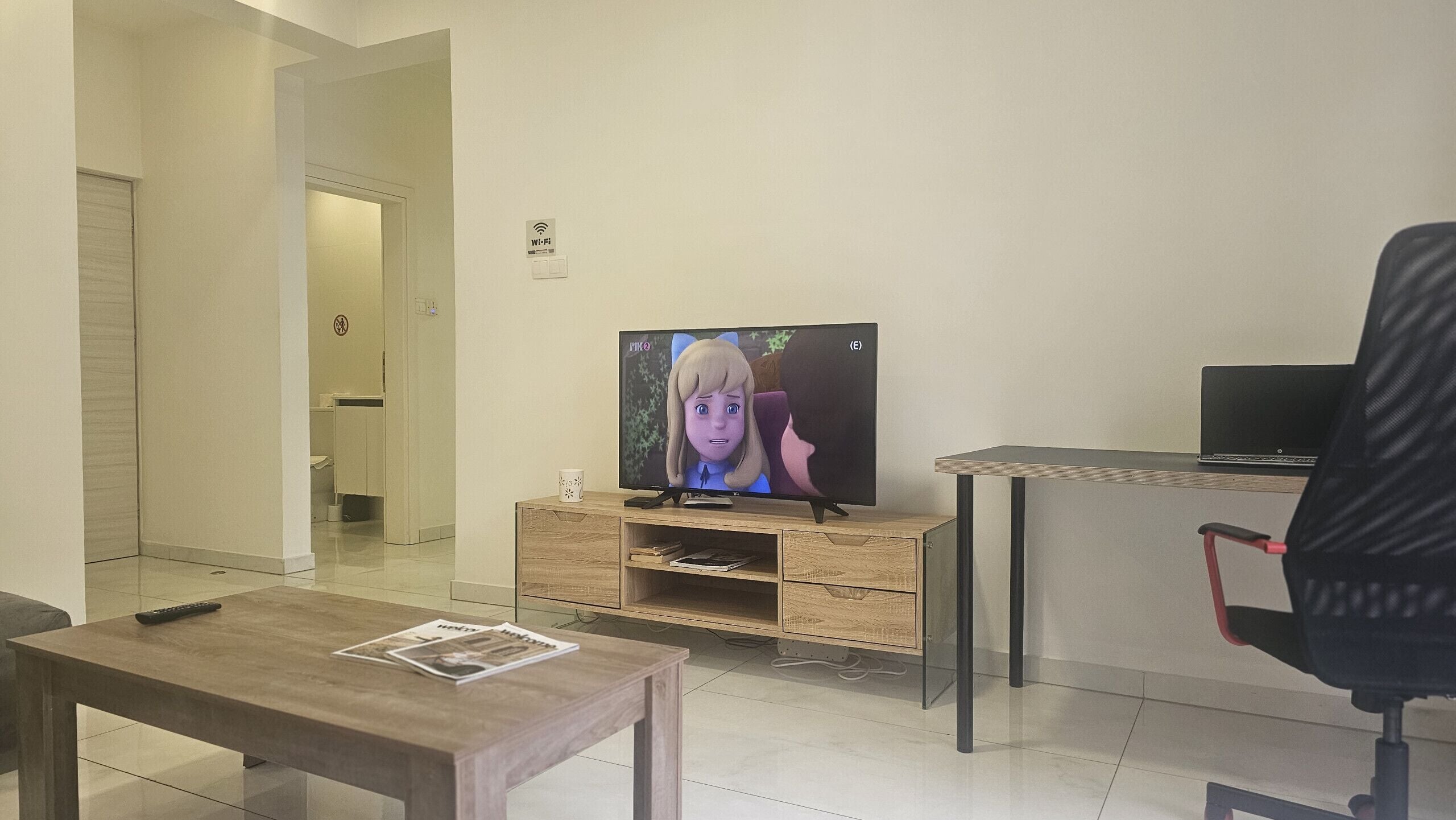 Una sala de estar moderna con un televisor de pantalla plana sobre un soporte de madera, una mesa de centro con un control remoto, una silla de oficina negra y un ordenador portátil sobre un escritorio.