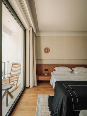 Habitación de hotel con una cama de madera, una mesita de noche, un reloj de pared, una silla y vistas a través de un ventanal amplio.