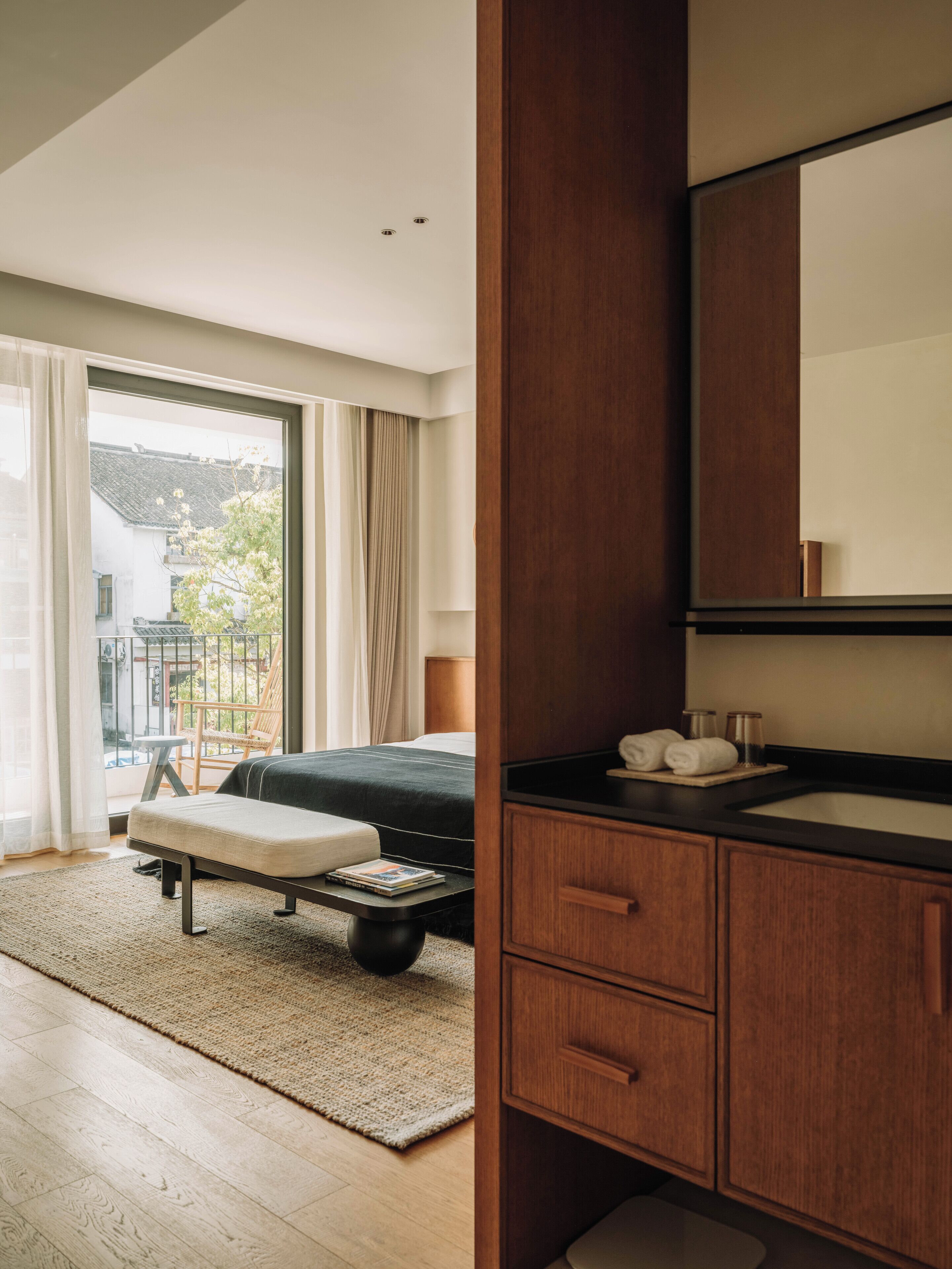 A modern hotel room with a bed, a small table with a magazine, a wooden bench, a large window with curtains, and a wooden vanity with a mirror and sink.