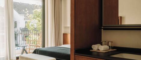 A modern hotel room with a bed, a small table with a magazine, a wooden bench, a large window with curtains, and a wooden vanity with a mirror and sink.