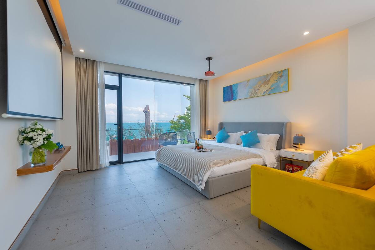 Un dormitorio moderno con una cama grande, un sillón amarillo, una mesita de noche y vistas al océano.