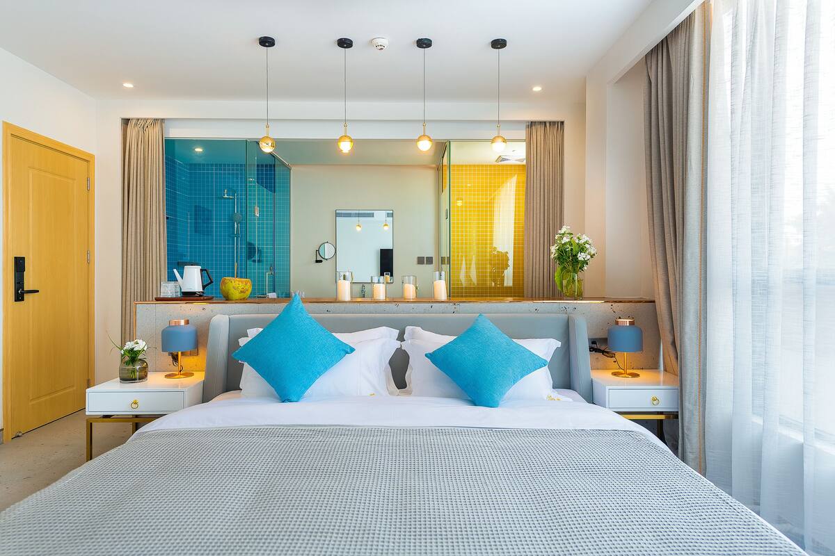 Una habitación de hotel moderna con una cama grande, dos mesitas de noche, un tocador, un espejo y vistas al baño.