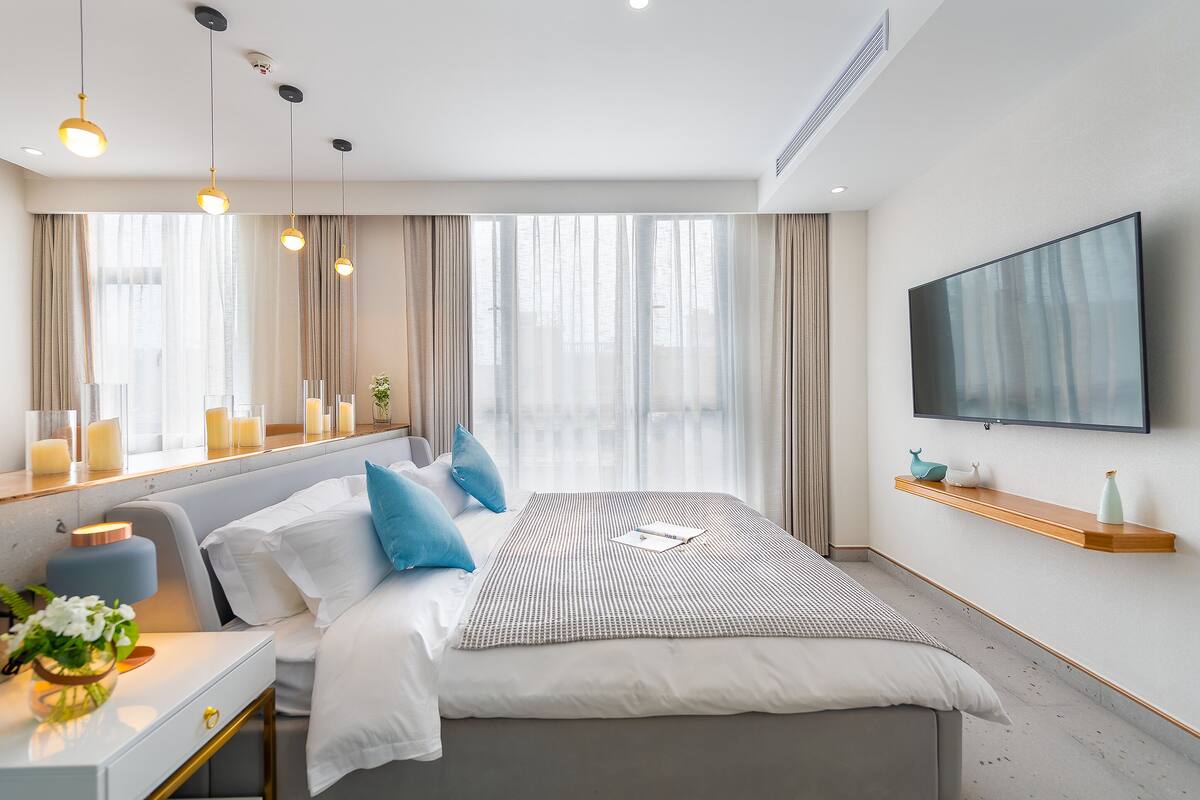 Un dormitorio moderno de hotel con una cama grande, lámparas de noche, un televisor y una ventana con cortinas.