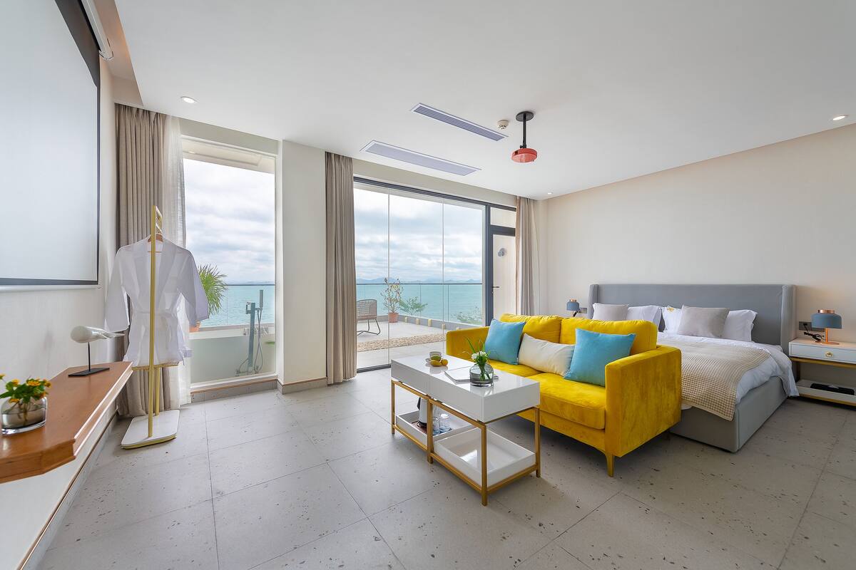 Una habitación de hotel moderna con una cama, un sofá amarillo, una mesa de centro blanca y un ventanal con vistas al mar.