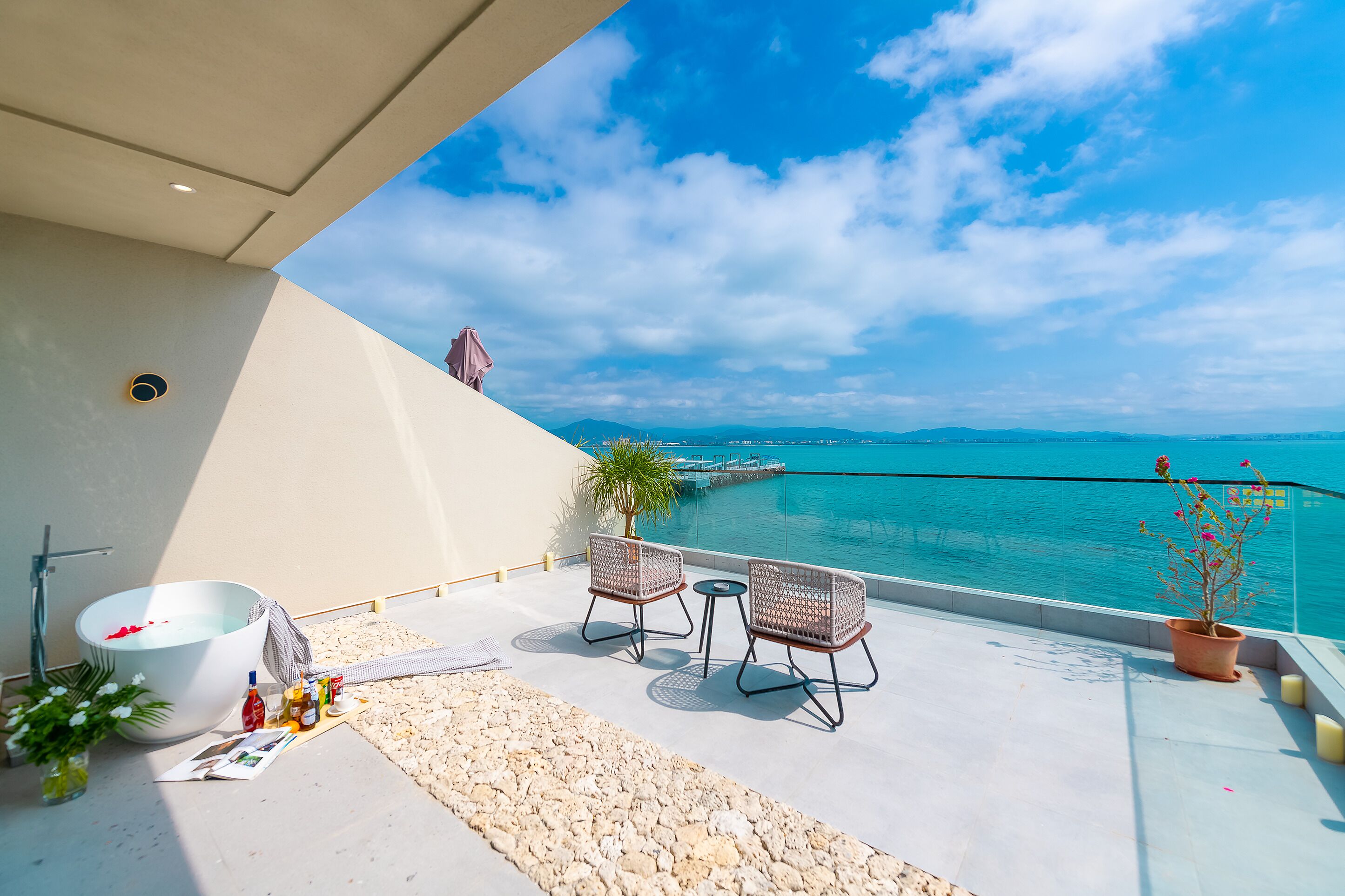 A balcony with a view of the sea, a bathtub, two wicker chairs, and a small table with bottles and a plant.