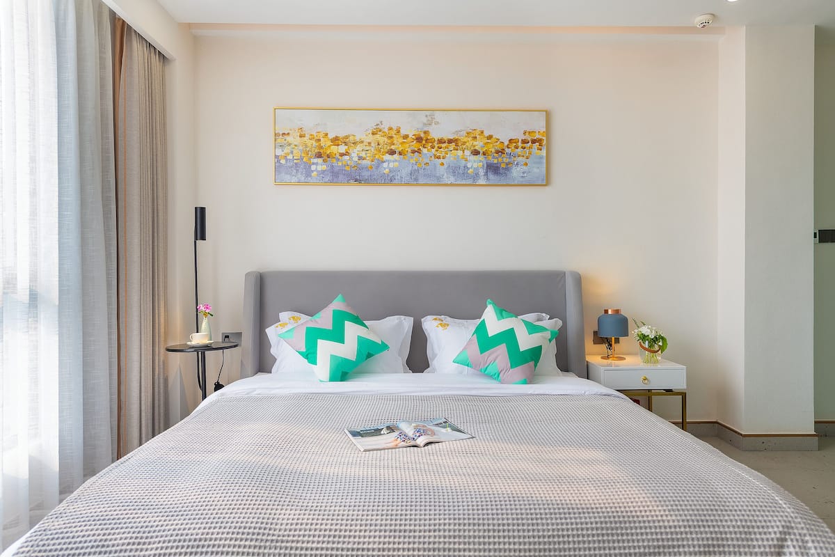 Habitación de hotel con una cama grande, dos almohadas, una mesita de noche con una lámpara, una pequeña mesa con un florero y un cuadro en la pared.