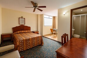 Hotelkamer met een bed, bureau, stoel, eettafel, plafondventilator, raam met gordijnen en een badkamer met toilet en douche.