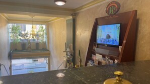 A hotel reception area with a television displaying a program, a reception desk, and a view of the outside through a glass door. - HOTEL MARIA CRISTINA (San Luis Potosí)