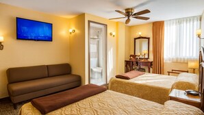 Une chambre d’hôtel équipée d’un canapé, de deux lits simples, d’un bureau avec une chaise, d’une télévision, d’un ventilateur de plafond et d’une salle de bain visible à travers une porte ouverte.