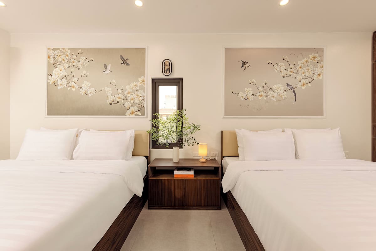 Una habitación de hotel con dos camas, una mesita de noche con lámpara, un jarrón con flores y dos cuadros colgados en la pared con motivos de aves y flores.