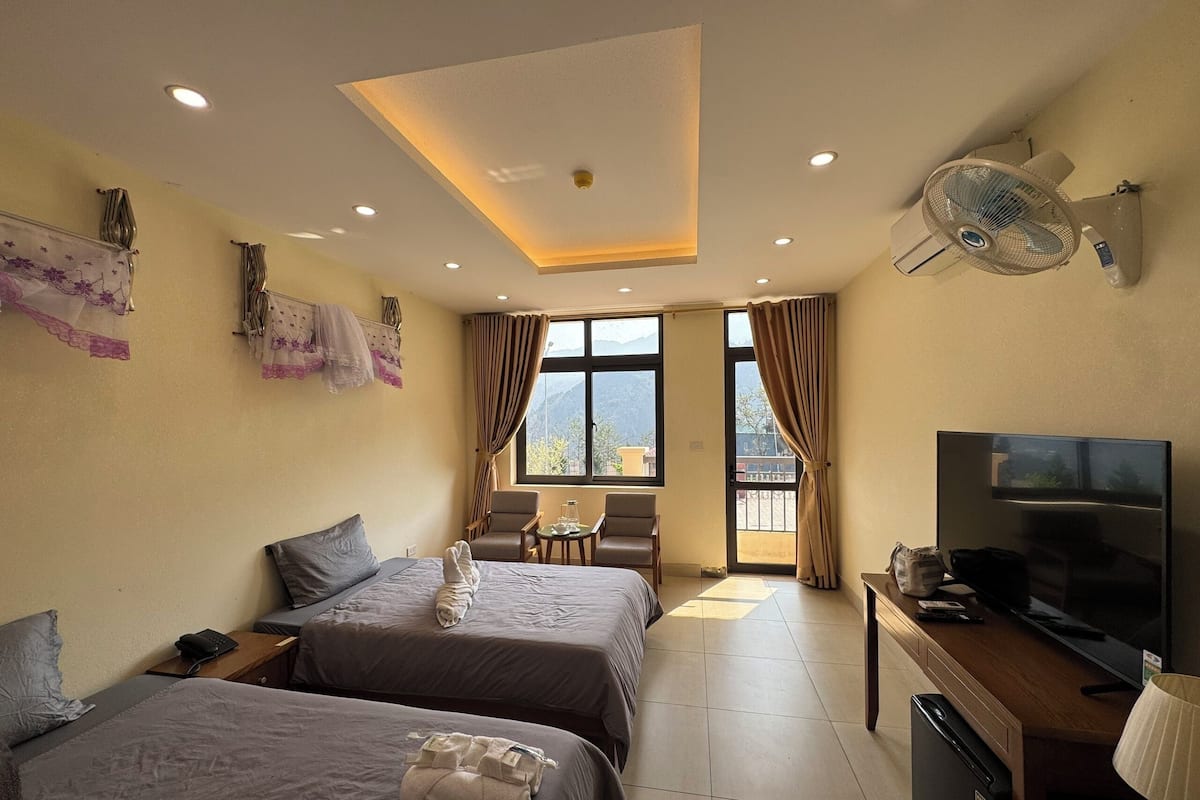 A hotel room with two beds, a desk with a TV, a fan, and a view of the mountains through the windows.