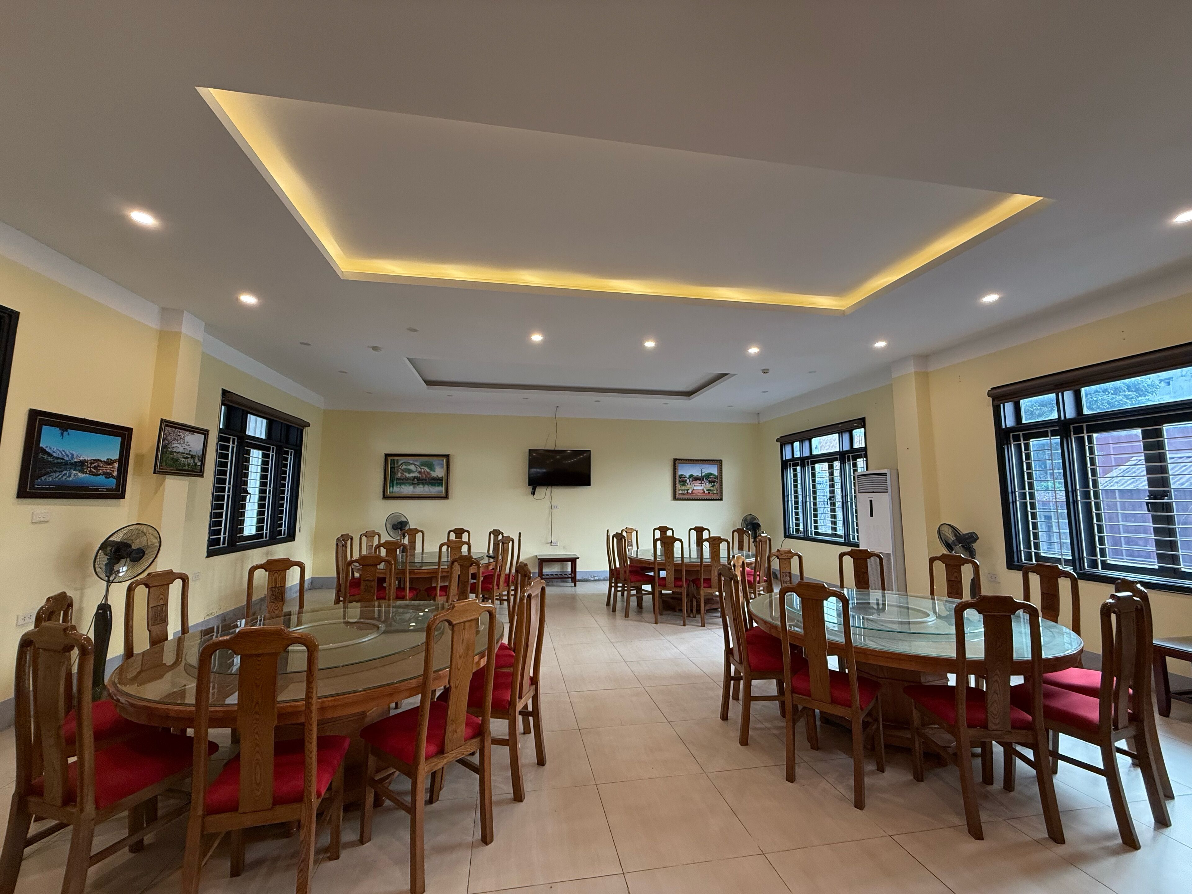 A dining area with round glass tables and wooden chairs, equipped with ceiling fans, and large windows with blinds.