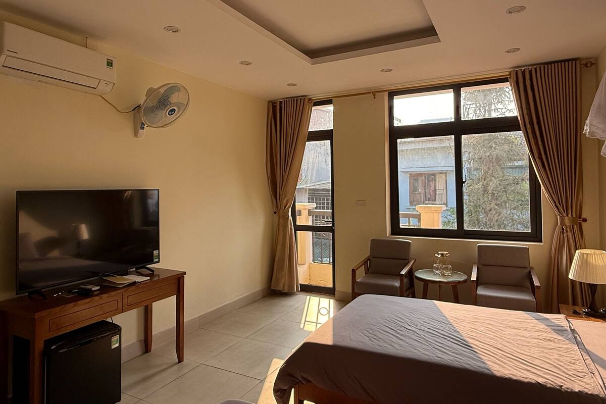 A hotel room with a bed, a TV, a desk, a refrigerator, a fan, a window with curtains, and a balcony.