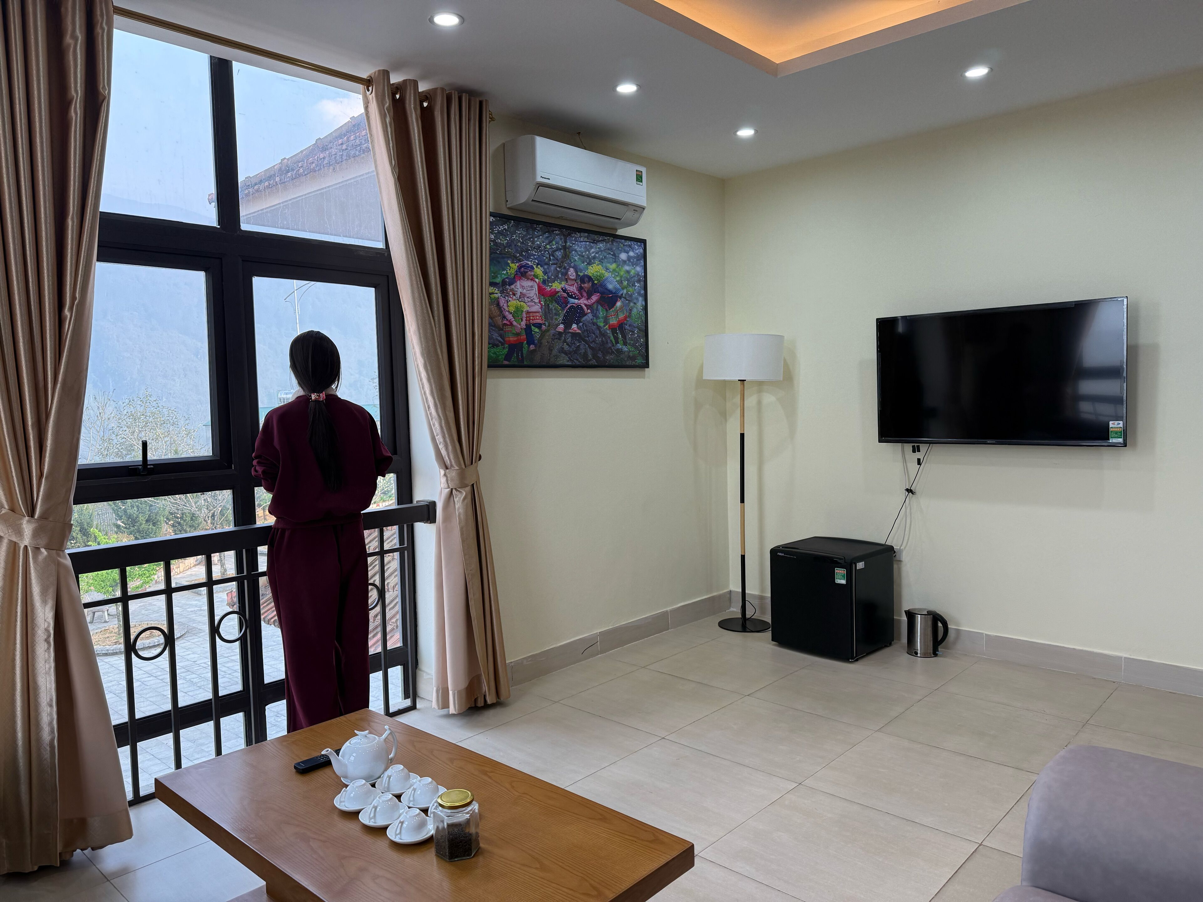 A hotel room with a large window, a flat-screen TV, a standing lamp, a coffee maker, and a table with tea sets.