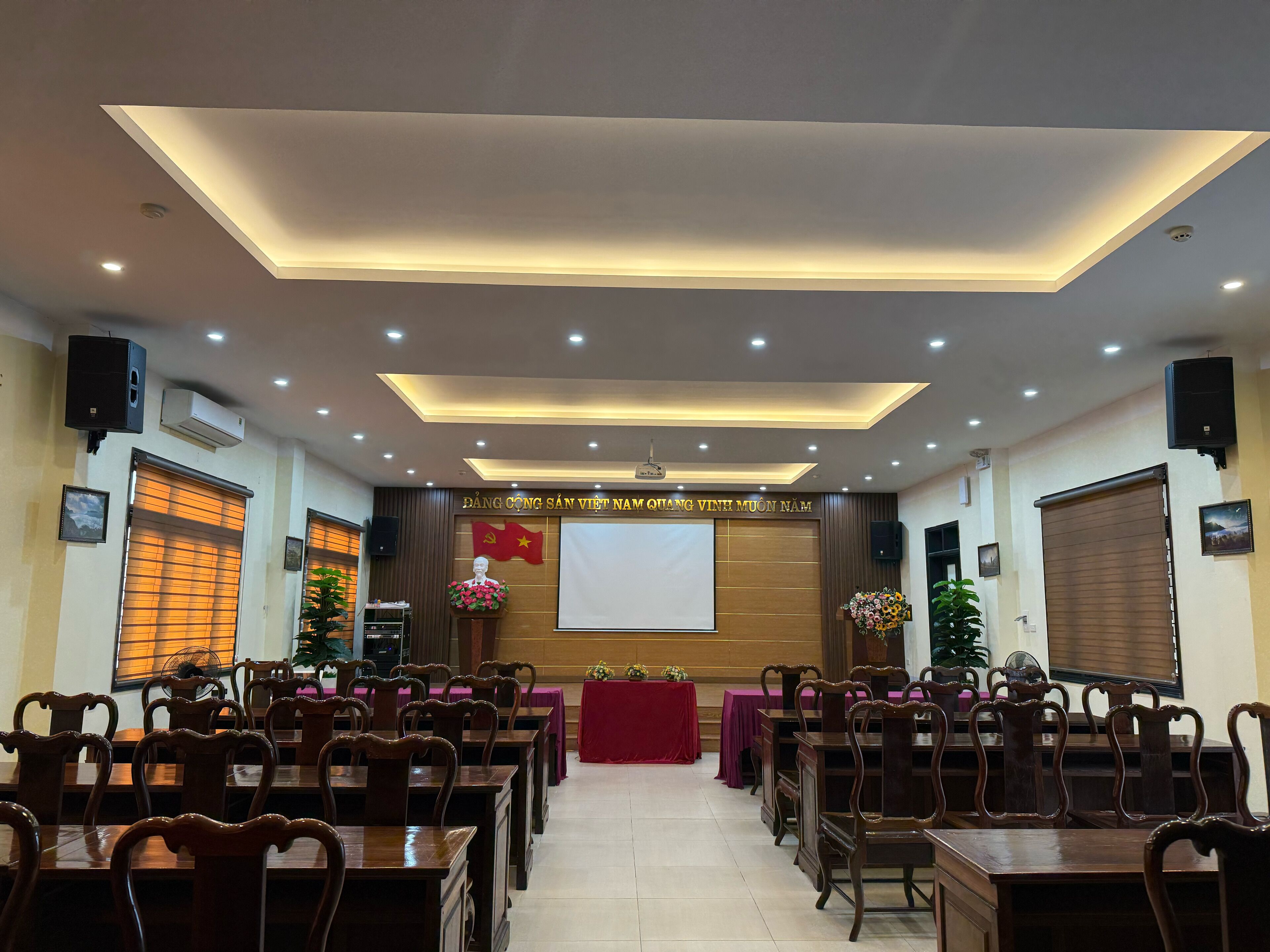 Un salón de conferencias con escenario, sillas de madera, mesas y un atril.