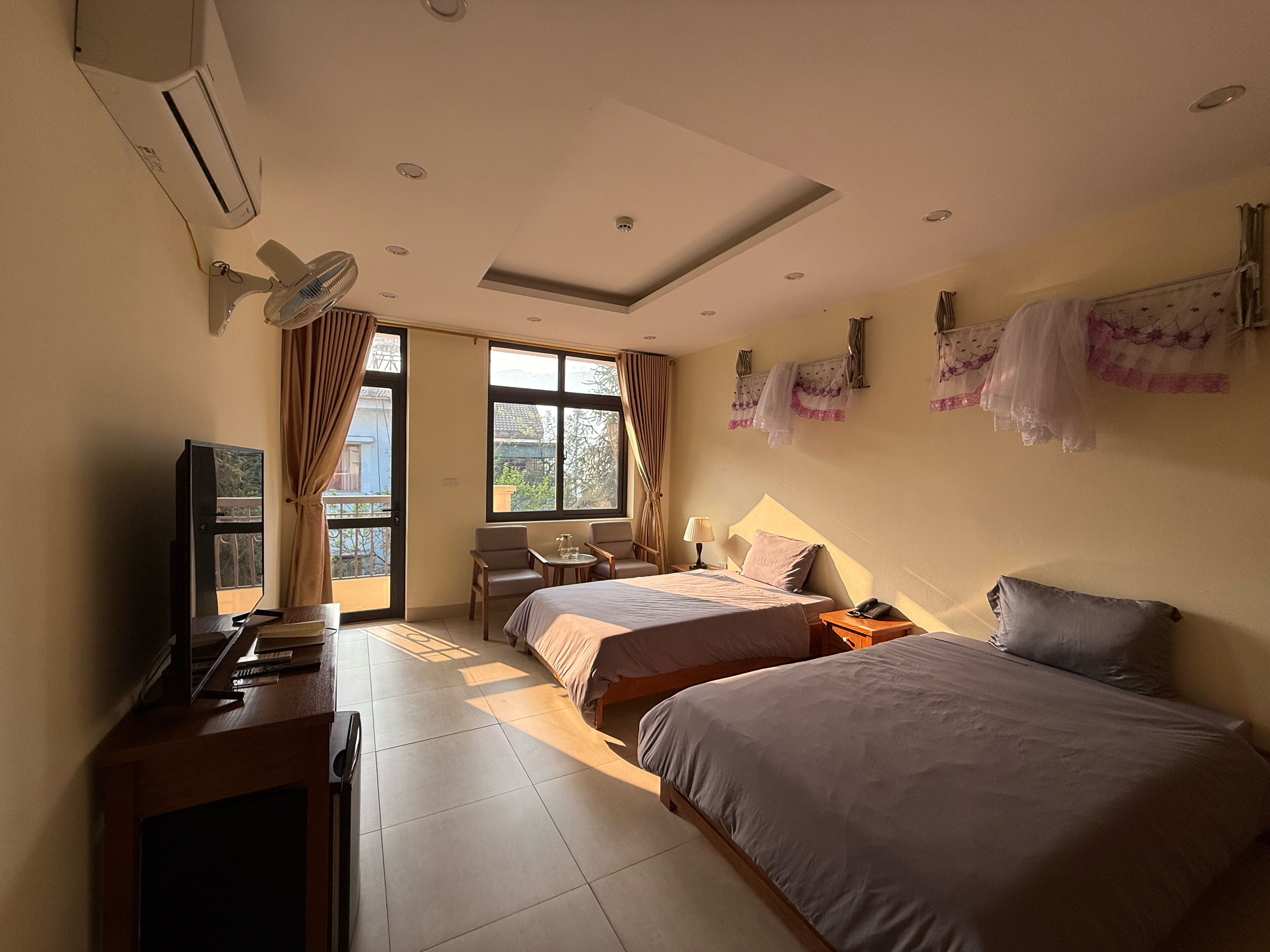 Quarto de hotel com duas camas, uma mesa de trabalho com televisão, uma cadeira, uma mesinha e uma janela com cortinas.