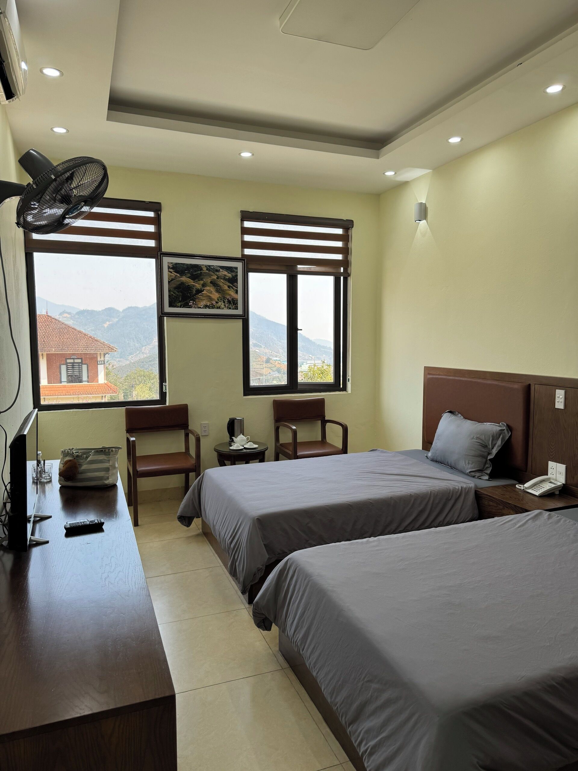 A hotel room with two beds, a desk with a TV, a fan, and a window with a view of mountains.