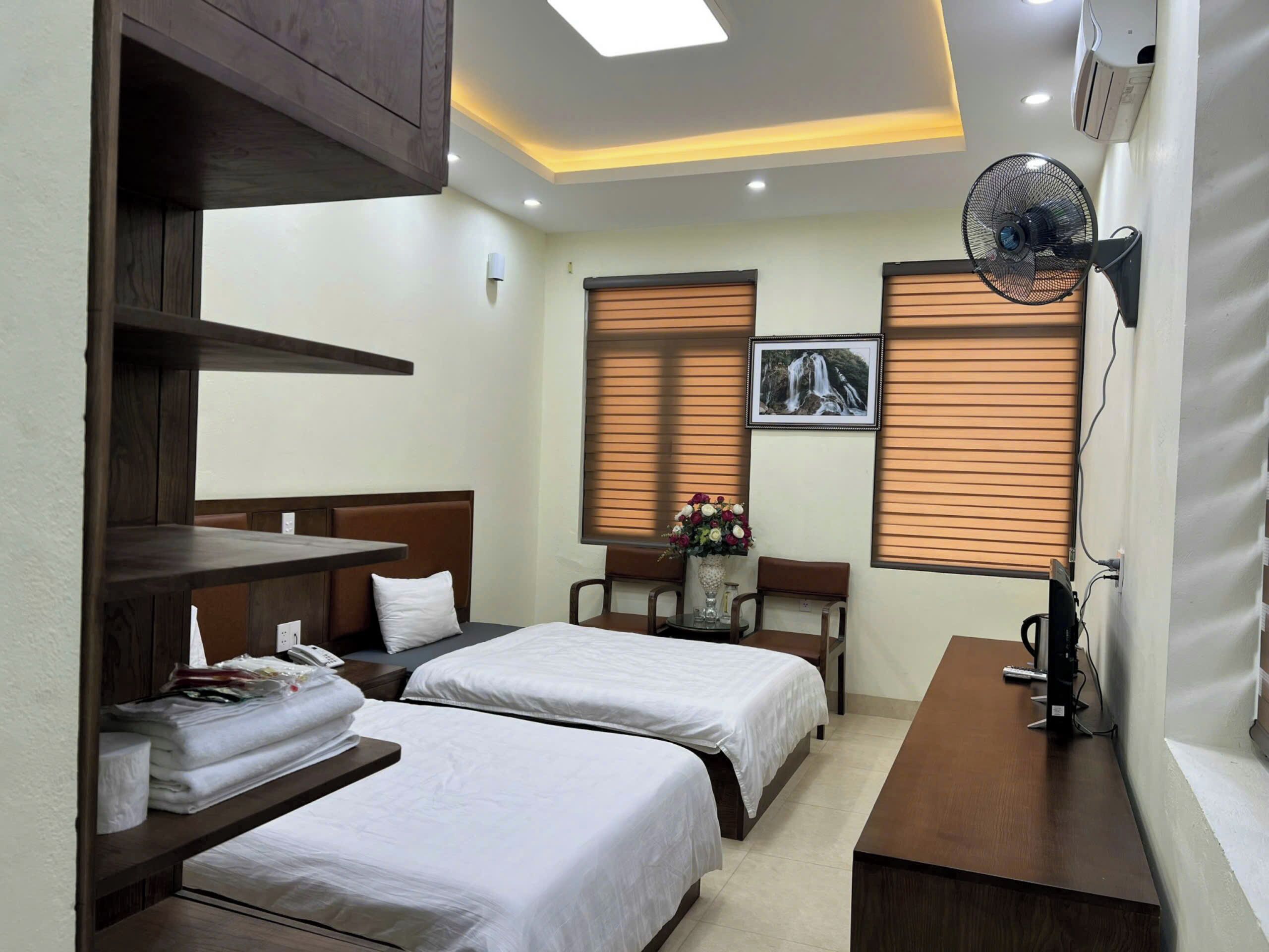 A hotel room with two beds, a desk, a chair, a fan, and a picture on the wall.