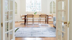 Una habitación amplia con suelo de madera, zona de comedor con mesa y sillas, y ventanas grandes que permiten la entrada de luz natural.